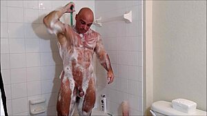 Muscular gay bodybuilder applies soap and oil to his body in the shower
