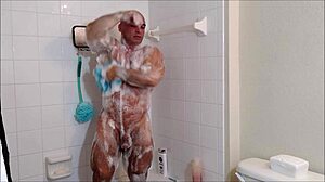 Muscular gay bodybuilder applies soap and oil to his body in the shower